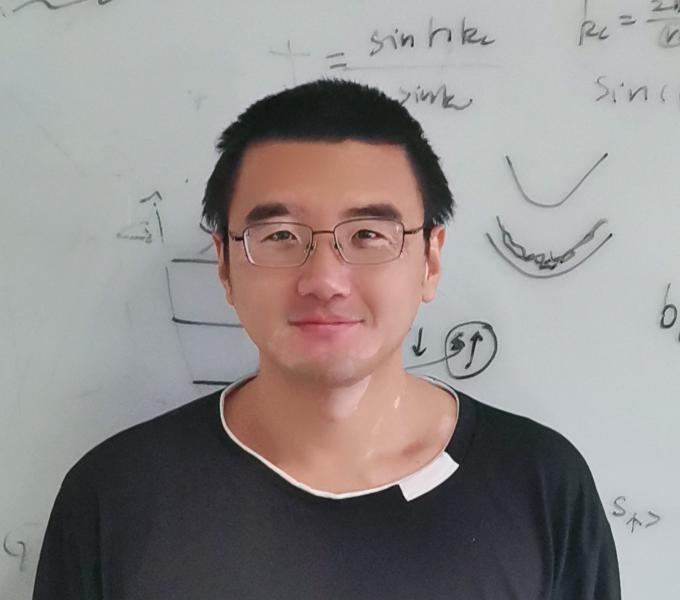 An image of Bao-Zong Wang, with a whiteboard behind him containing physics notes.
